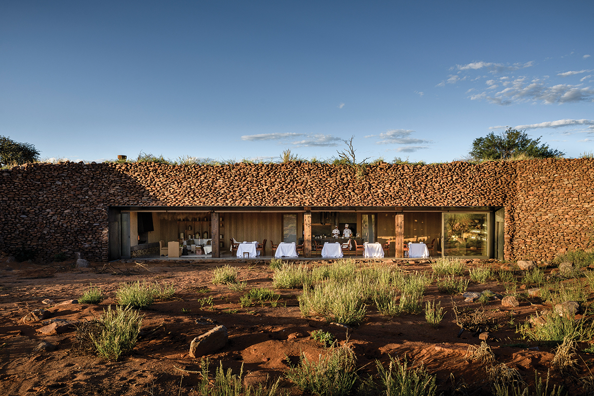 A stone-walled, farmhouse restaurant in a remote desert landscape. Large glass windows reveal the interior, where tables are set for dining.